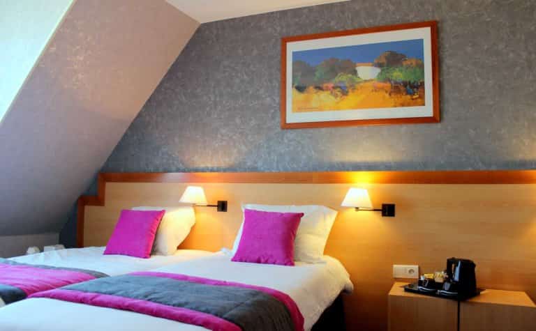 Chambre avec deux lits jumeaux décorés de coussins et plaids violets, appliques murales éclairant une tête de lit en bois, et tableau coloré au mur.